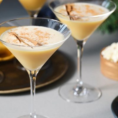 A trio of indulgent Eggnog Martini cocktails presented in long-stemmed Martini glasses, surrounded by snack bowls on a light grey surface