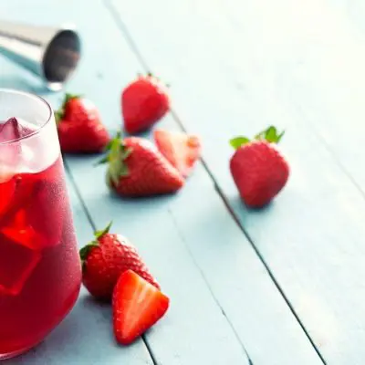 Close up of two Strawberry shrub cocktails in tumbler gasses, garnished with fresh strawberries, presented on a rustic, light blue surface