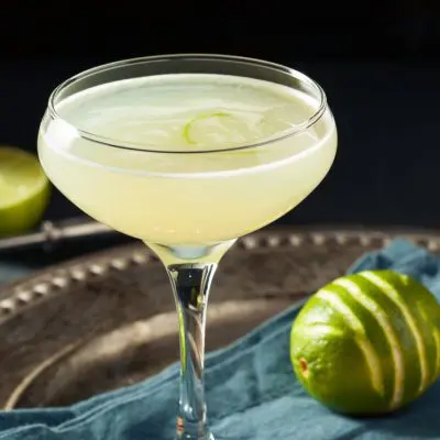 A transluscent Pegu Club cocktail on a pewter serving platter along with a scored lime, against a dark backdrop