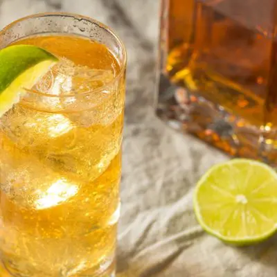 Close-up top shot of a Bourbon Rickey garnished with a piece of lime, with a decanter of bourbon and a sliced lime visible the side