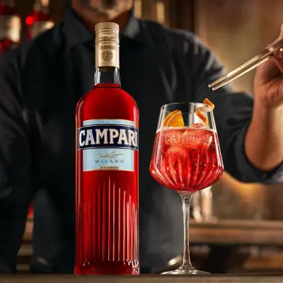 A close up of a bottle of Campari with a bartender adding garnish to a Campari Spritz cocktail