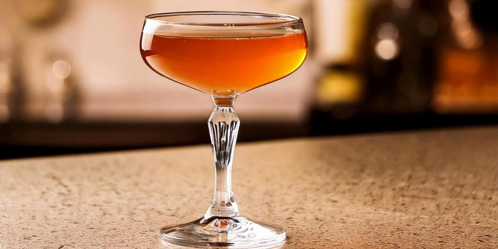 A coupe glass with a rich orange-hued cocktail, set on a sleek bar counter. The lighting highlights the clarity of the drink and the elegant detailing on the glass stem.