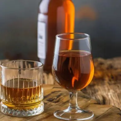 Two glasses of rye whiskey and a snifter glass of bourbon set on a rustic wooden surface against a grey backdrop