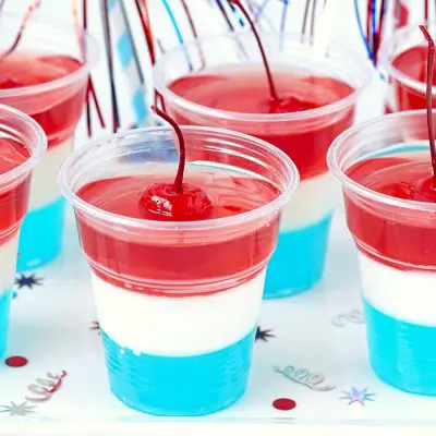 Close up of a collection of blue, white and red layered jello shots in plastic shot glasses, garnished with red cherries