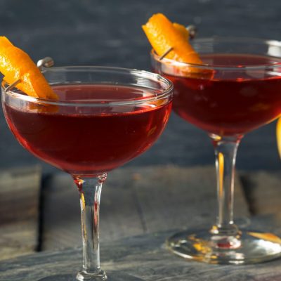 Two bourbon-based Revolver Cocktails garnished with flamed orange twists against a pewter backdrop with a peeled orange and a container of bourbon in the background