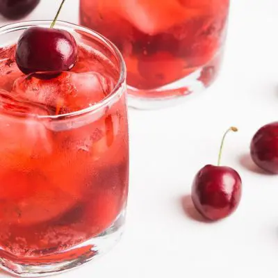 A pretty pair of red Dirty Shirley cocktails garnished with fresh cherries, posed on a white surface