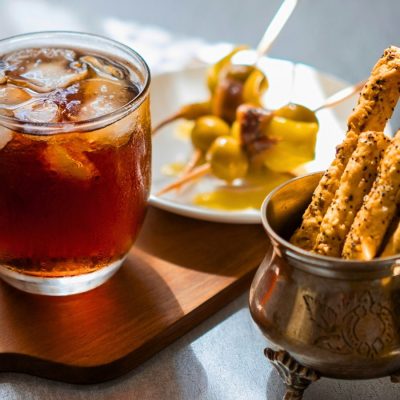 Close up view of a Cynar Spritz cocktail served on a wooden platter with a variety of snacks, presented on a white linen surface