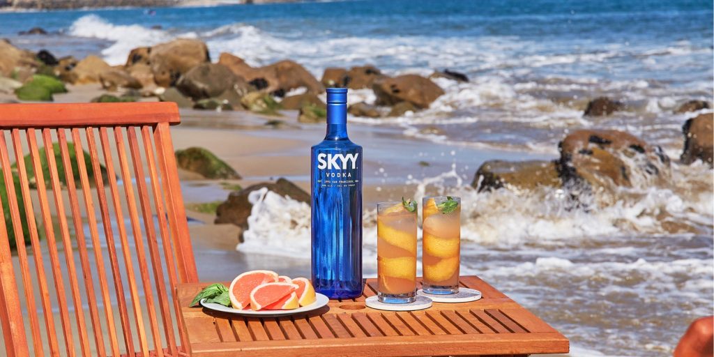 Two Turtle Dove SKYY rainbow cocktails served on a wooden table with a bottle of vodka and a plate of grapefruit wedges, in a beach setting