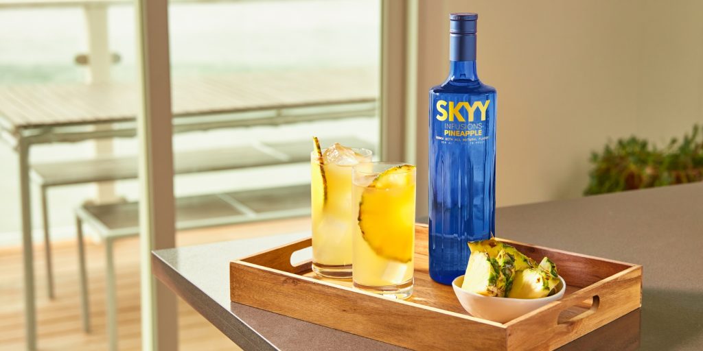 Two Ancho Pineapple Sodas served on a wooden tray with a bottle of SKYY vodka and bowl of pineapple wedges on the side