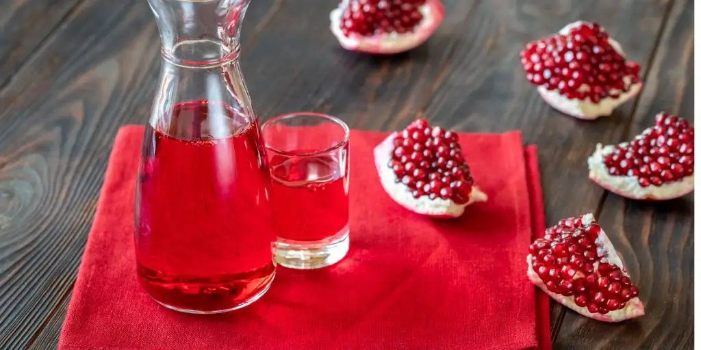 Close up of pomegranate syrup in a decanter, next to a shot glass of grenadine and quartered fresh pomegranates on a red linen napkin