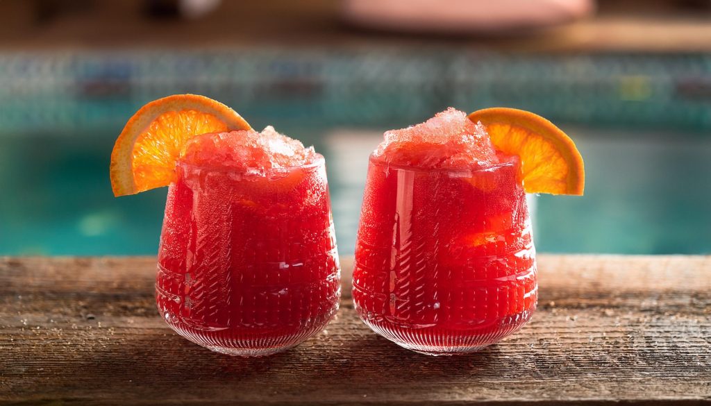 Close-up of two bright red Frozen Negroni cocktails with orange garnish, served poolside