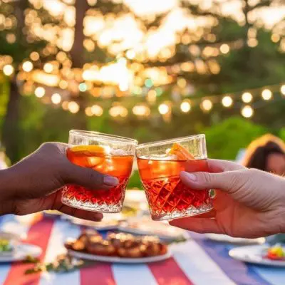 Two friends clinking Negroni cocktails at a Memorial Day picnic with a vibrant crowd in the background at dusk