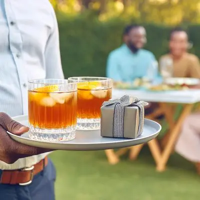 A man holding a tray of two bourbon cocktails for Father's day and a gift, people enjoying lunch in the background