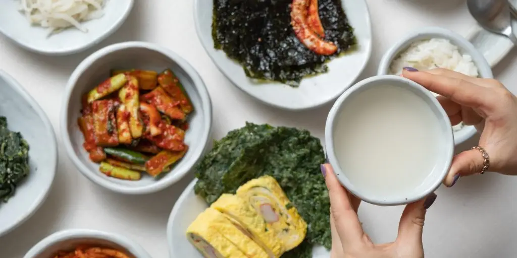 Top shot of a woman holding a cup of Makgeolli over a table filled with delicious Korean food