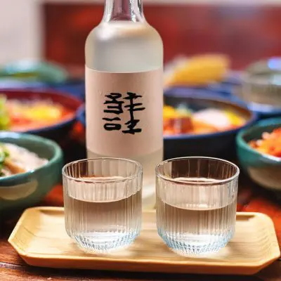 Two sipping glasses of clear spirit on a table next to a bottle of Korean spirit set on a table filled with Korean food