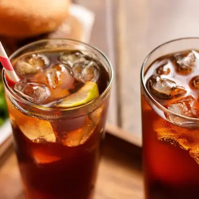 Two White Rum cocktails with cola in tall glasses next to a burger on a wooden table