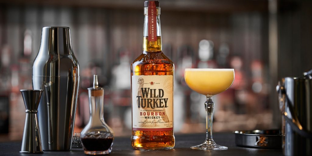 Close up of a Whiskey Sour in a coupe glass next to a bottle of Wild Turkey Bourbon on the counter of a modern bar in daytime