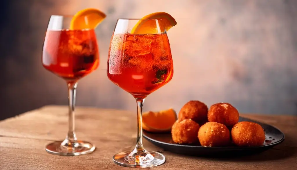 Two Campari Spritz cocktails with orange garnish, served with a plate of arancini