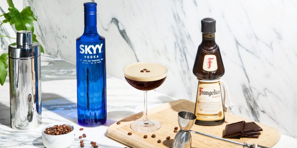 A Hazelnut Espresso Martini with a foamy top, garnished with three coffee beans, next to a blue bottle of SKYY Vodka and a bottle of Frangelico, cream and white themed background and props