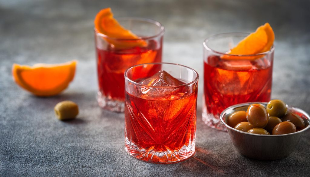 Close up of three French Negroni cocktails, garnished with an orange twist each, in rocks glasses on a slate grey surface