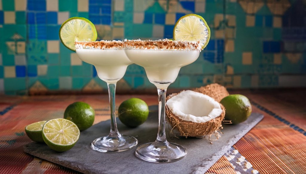 Two Coconut Margarita cocktails with taoasted coconut rims on a table in a modern living room against a blue patterned backdrop, surrounded by fresh limes and coconuts