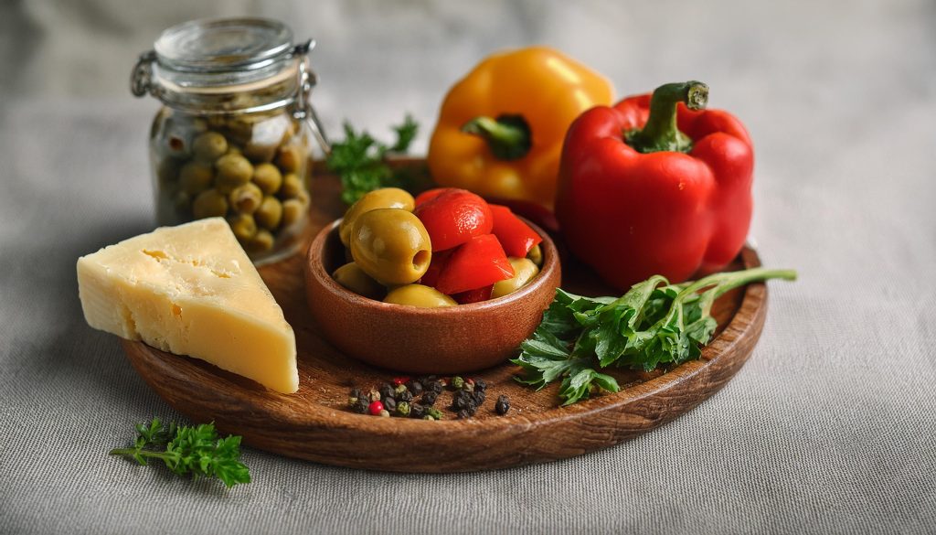 Close up of a wooden board with peppers, herbs, a jar of green olives and a wedge of parmesan cheese