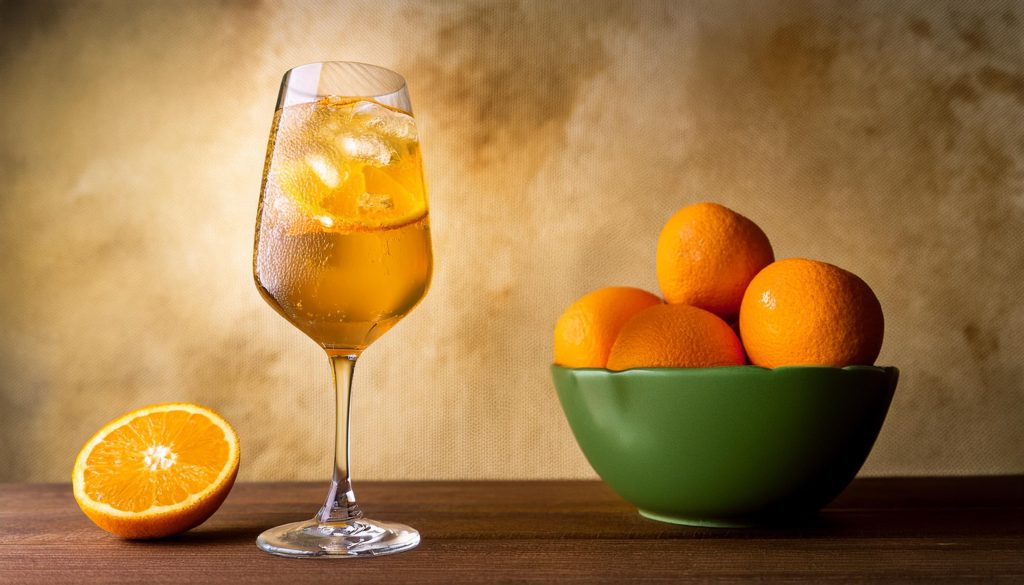 Close up of a Citrus Spritz next to an olive green bowl of orange, against a background in different shades of brown