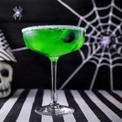 A ghoulish green Beetlejuice Margarita on a striped tablecloth against a spooky Halloween backdrop