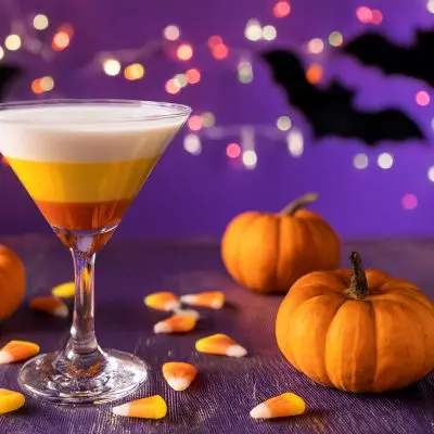 A layered Candy Corn Martini, served in a Halloween setting with purple background with fairy lights