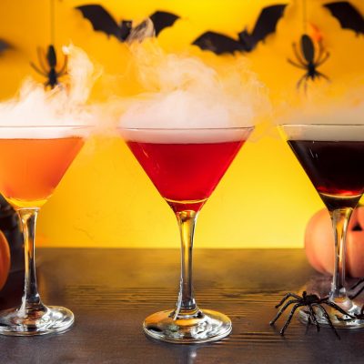 Three SKYY Vodka Halloween Martini cocktails in orange, red and black, set against a yellow background with spiders hanging from the ceiling and bats on the wall