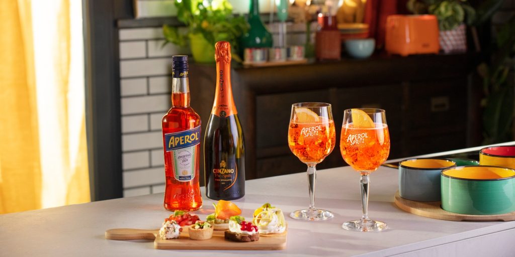 A styled shot of two vibrant orange Aperol Spritz cocktails next to a bottle of Aperol and Cinzano Prosecco