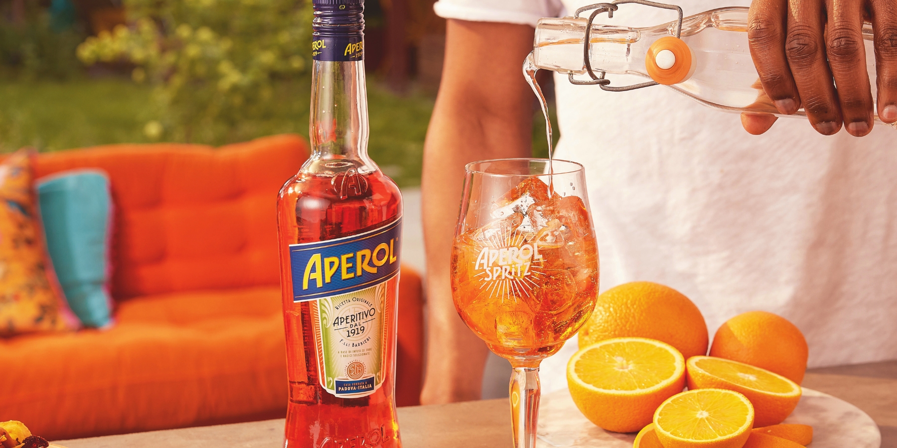 A young man pouring club soda into his Aperol Spritz, vibrant orange couch in the background