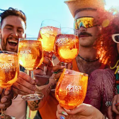 Close-up of friends outdoors in the sunshine clinking together their vibrant orange Aperol Spritz cocktails