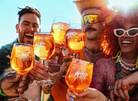 Close-up of friends outdoors in the sunshine clinking together their vibrant orange Aperol Spritz cocktails