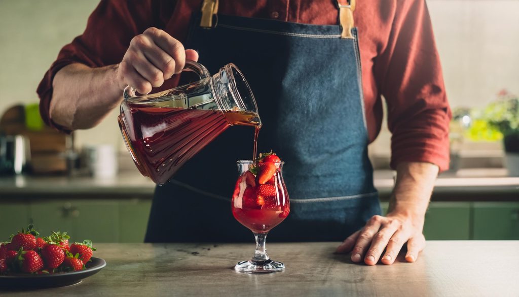 A man in an apron pouring a homemade berry shrub mixer into a strawberry cocktail