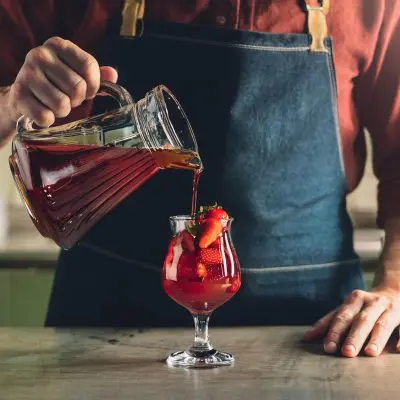 A man in an apron pouring a homemade berry shrub mixer into a strawberry cocktail