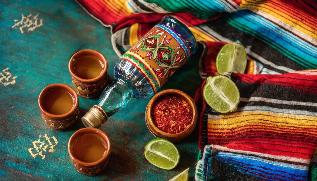 Top view of a selection of gifts for requila lovers on a colorful fabric backdrop, including a bottle of tequila, interesting salts and more