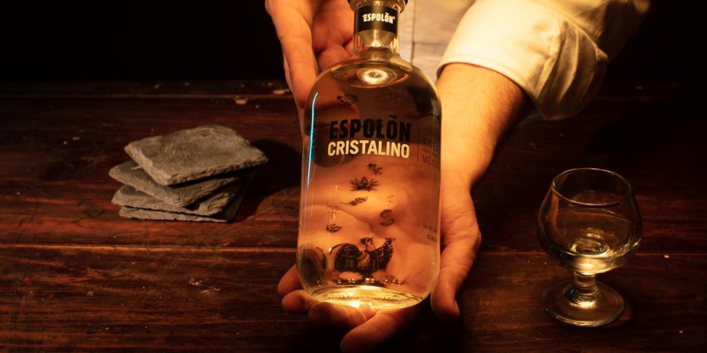 Close up of a man's hand holding a bottle of Espolon tequila over a dark wooden counter