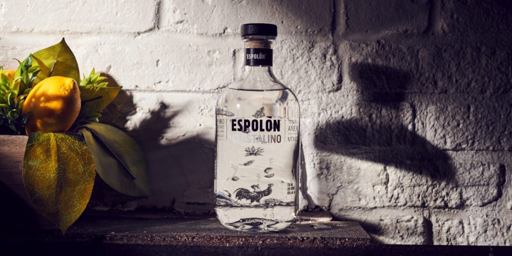 A close-up of a bottle of Espolòn Cristalino Tequila against an off white brick wall with a bowl of lemons to the side