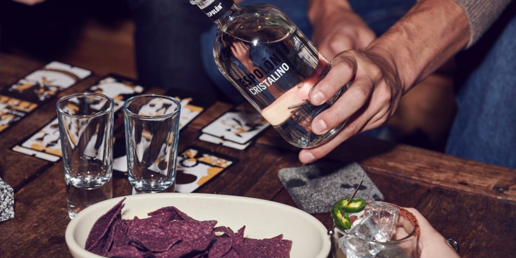 A close-up shot of a hand that's about to pour Espolòn Cristalino tequila into a glass on a table with playing cards and beetroot chiips