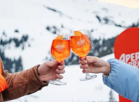 Two hands clinking together bright orange Aperol cocktails in an Apres Ski setting, snowboard and snowy mountains in the background