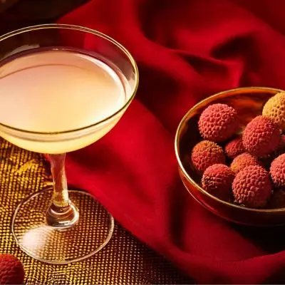 A delicate cocktail in a coupe glass accompanied by a bowl of fresh lychees, set against a rich red fabric and golden accents.