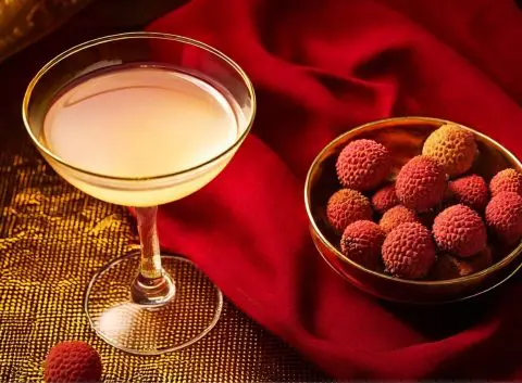 A delicate cocktail in a coupe glass accompanied by a bowl of fresh lychees, set against a rich red fabric and golden accents.