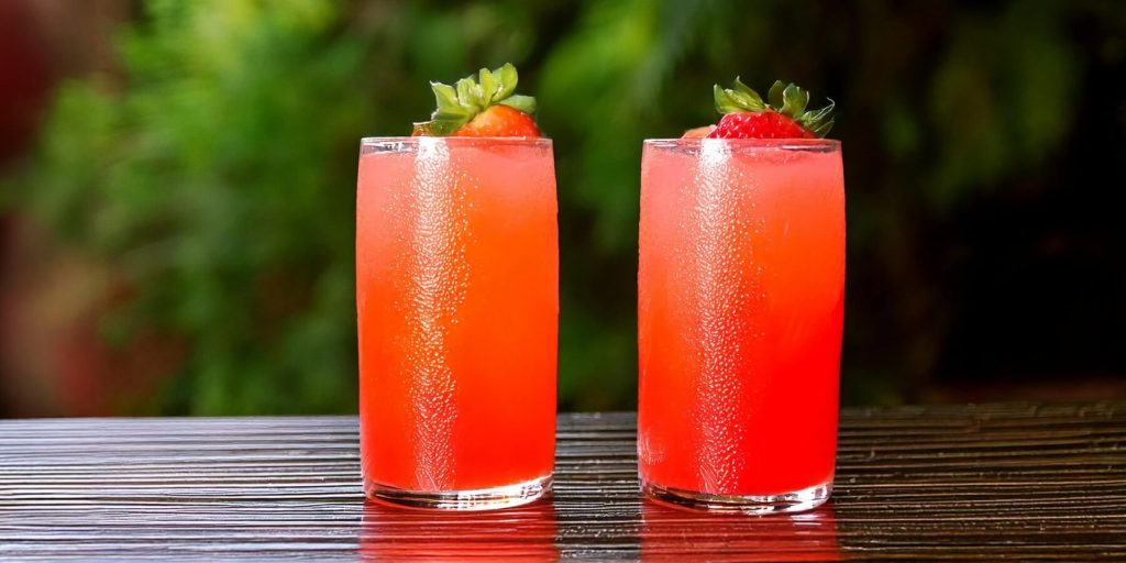 Two tall glasses of bright red Strawberry Highball cocktails garnished with fresh strawberries, set on a shiny, wet surface with lush greenery in the background.