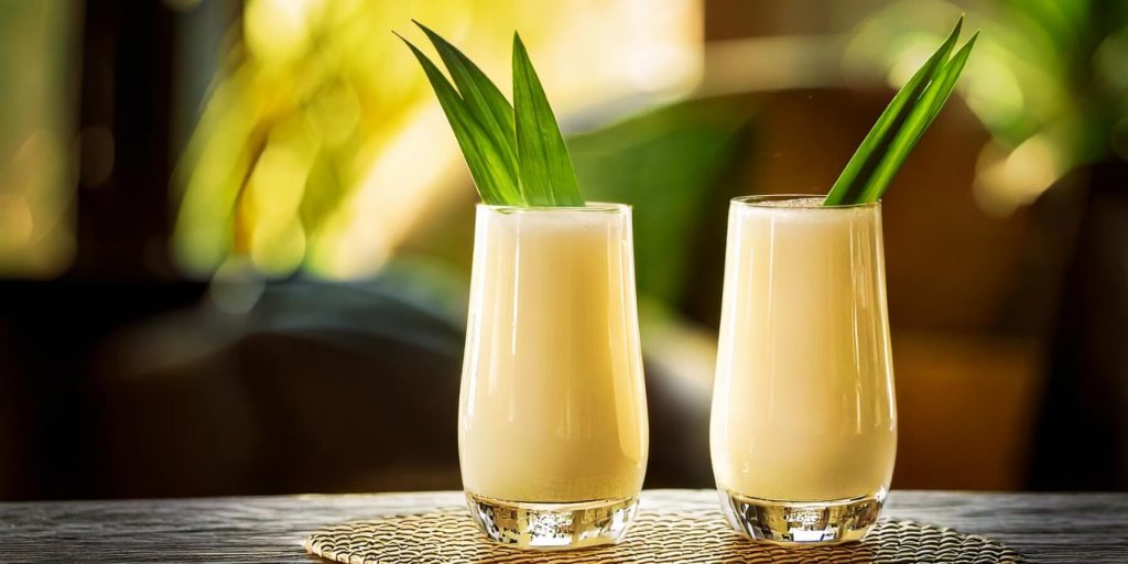 Two creamy, pale yellow coladas served in tall, curvy glasses, each garnished with pandan leaves, on a table with a summery background.