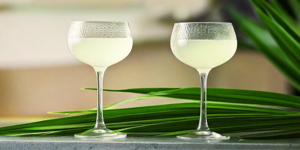 Two frosty, pale green Gimlets served in coupe glasses, placed on a light gray surface surrounded by fresh pandan leaves.