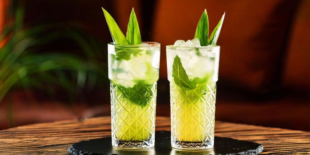 Two refreshing green Mojitos garnished with mint leaves and pandan leaves, served in tall crystal glasses filled with ice, on a wooden table.