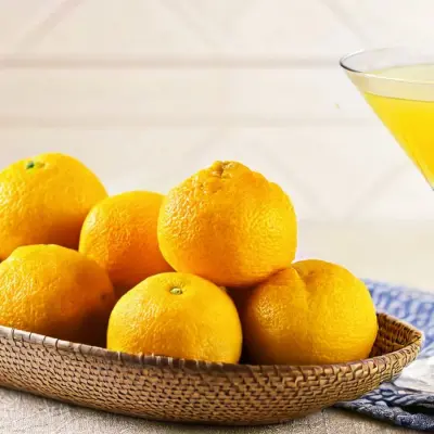 A vibrant display of fresh yuzu fruits in a woven basket sits beside a delicate coupe glass filled with a golden cocktail. A soft blue cloth napkin rests nearby, emphasizing the fresh, citrusy theme.