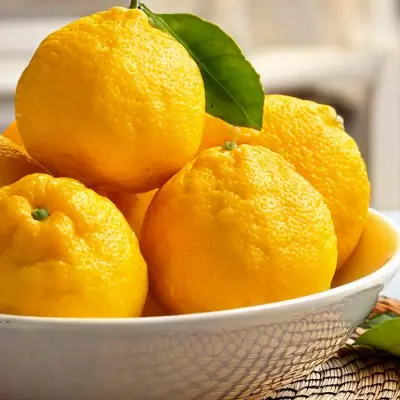 A ceramic bowl holds a pile of fresh, vibrant yellow yuzu fruits with their leaves still attached. The bright citrus tones contrast beautifully against the natural woven mat and soft background.