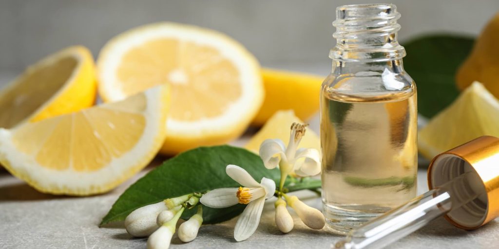 A small glass bottle filled with pale golden liquid, likely yuzu essential oil, sits next to a dropper with a gold cap. Surrounding it are fresh lemon slices, green leaves, and delicate white citrus blossoms. 